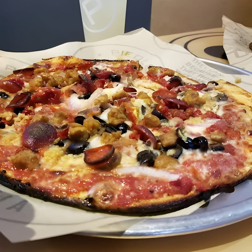 A close-up of a Hawaiian BBQ pizza with pepperoni, sausage, olives, and cheese on a metal tray.