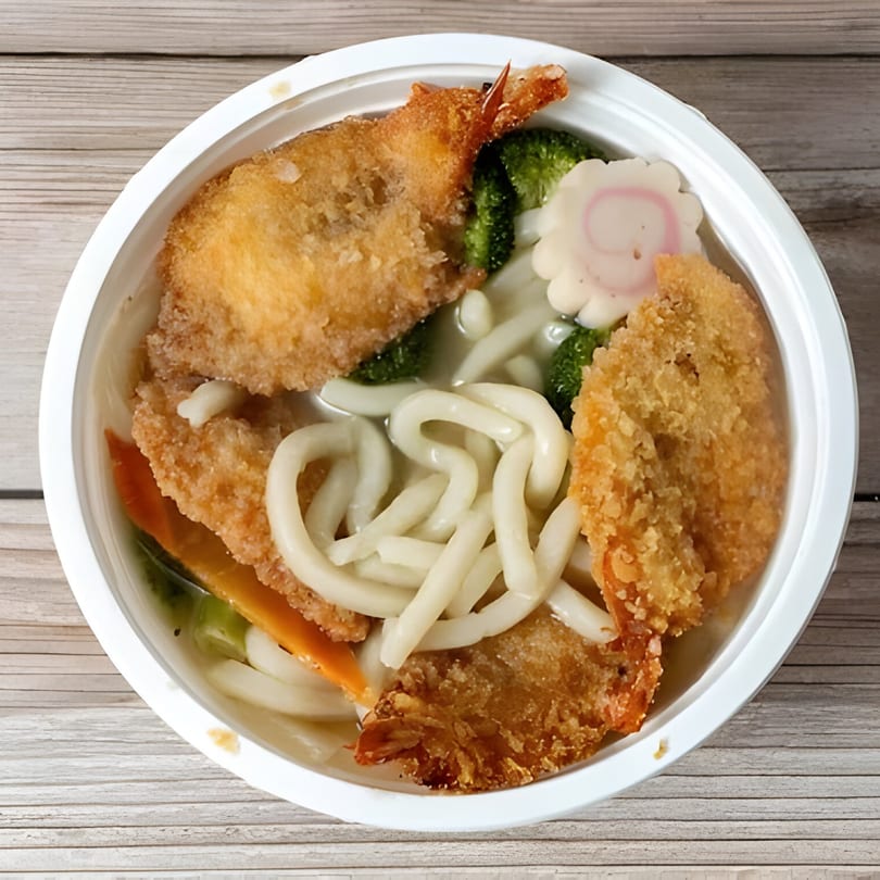 A white bowl of udon noodles with tempura shrimp, broccoli, carrots, and a fish cake.